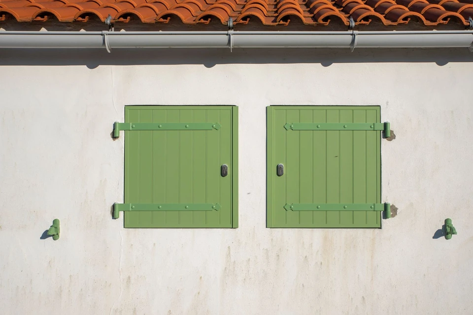 Volets verts fermés sur un mur blanc, sous un rang de tuiles